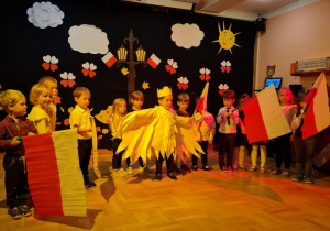 Dzieci z chorągiewkami biało-czerwonymi przedstawiają program artystyczny z okazji Narodowego Święta Niepodległości. W środku stoi chłopiec przebrany za Orła Białego.