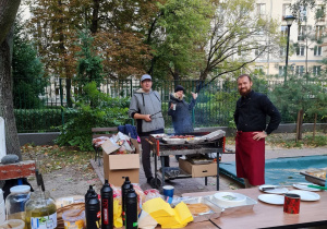 Rodzice grilują kiełbaski i kaszankę, a na stole naczynia, przyprawy i dodatki do grila.